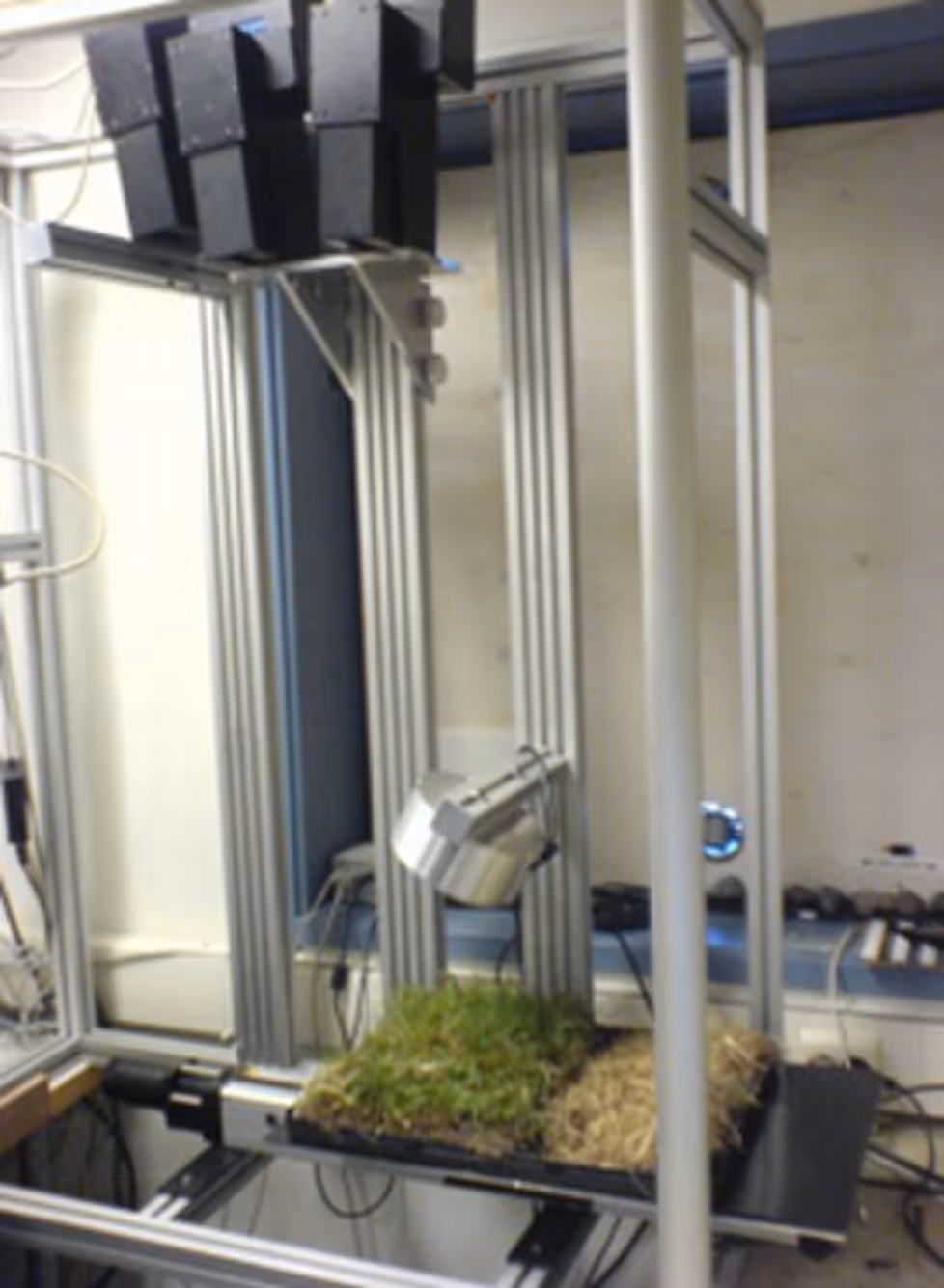 Laboratory setup with a metal frame supporting lights and electronic equipment over a tray containing soil and grass samples.