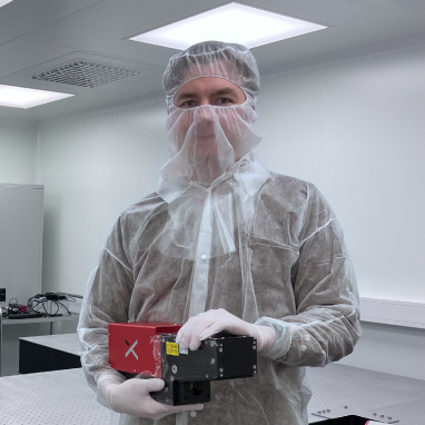 Researcher in cleanroom attire holding a red and black electronic hyperspectral HySpex camera in a lab.