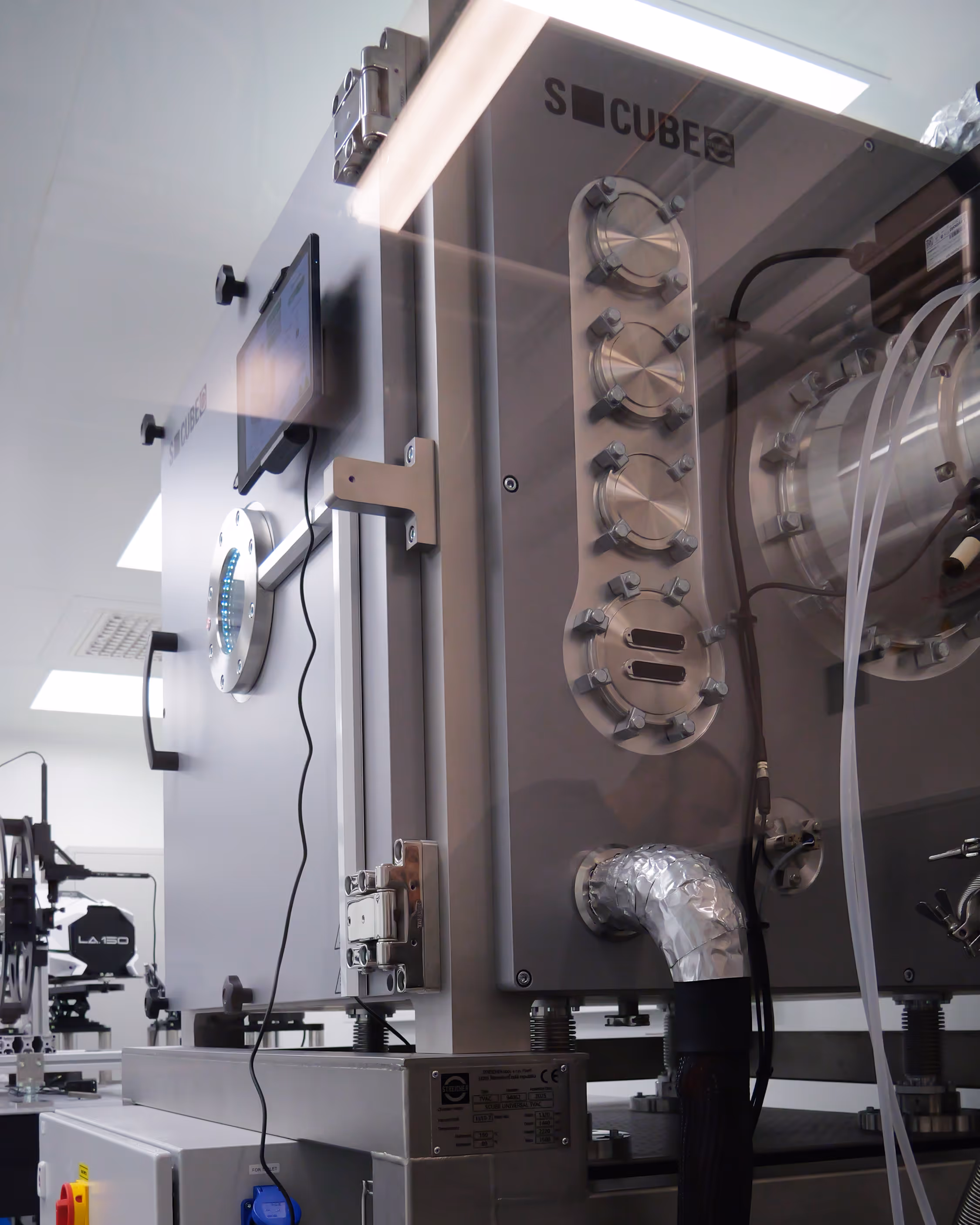 Thermal vacuum chamber labeled S-CUBE inside a cleanroom with pipes and control panel in a lab setting.