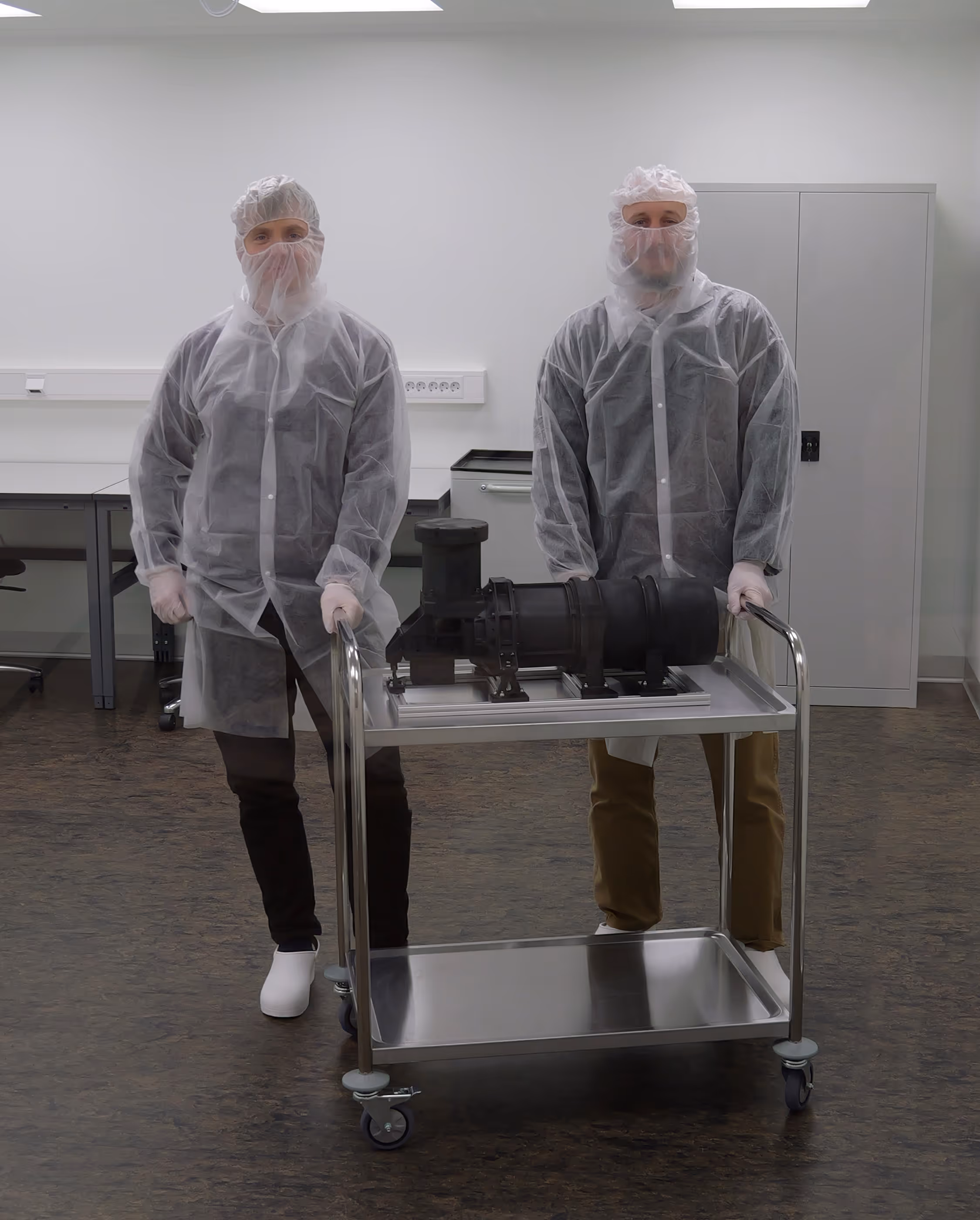 Two researchers in cleanroom suits standing next to a cart with a model of a HyperNOR HySpex hyperspectral camera in a sterile room.