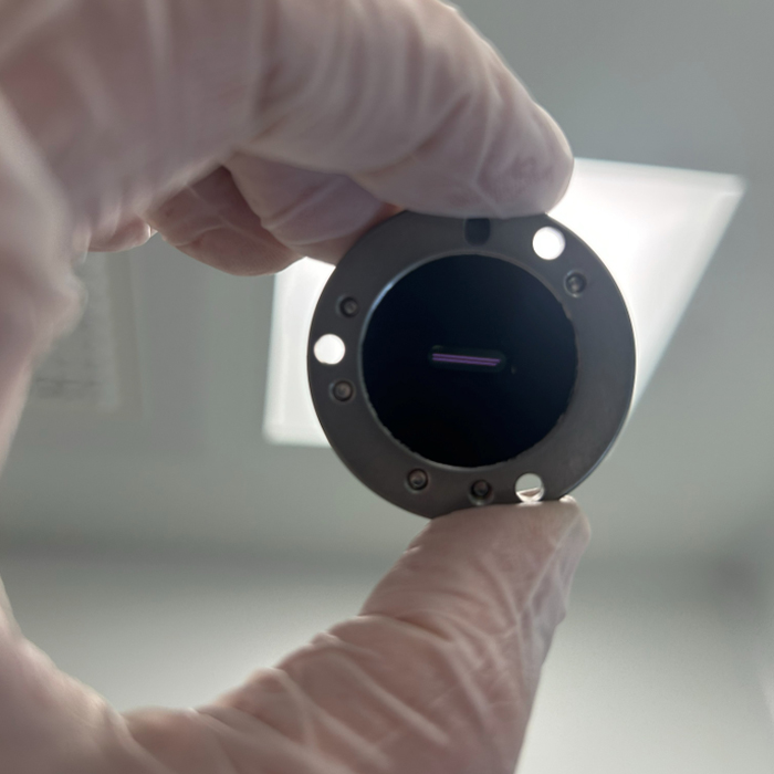 Hand in disposable glove holding a small circular camera lens with a dark center.