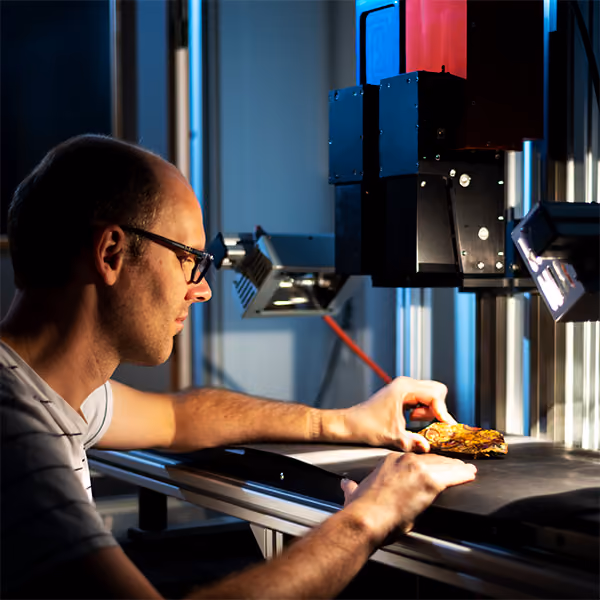 Man with glasses places a rock on a scanning stage underneath two HySpex Classic Series hyperspectral cameras for scanning purposes in a dimly lit lab setting.