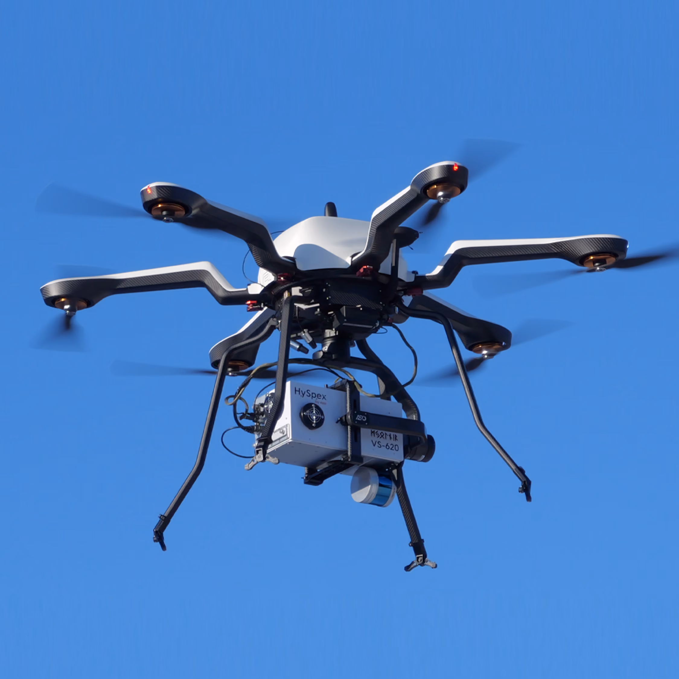 White and black hexacopter UAV drone with a HySpex Mjolnir VS-620 hyperspectral camera flying against clear blue sky