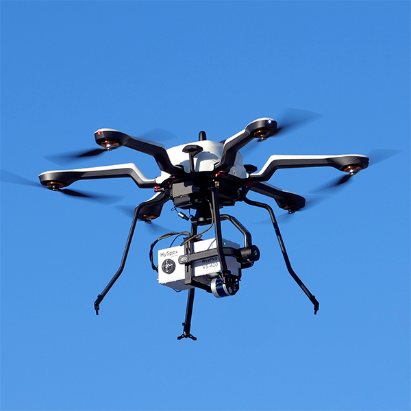 A white hexacopter UAV drone flying through a blue sky, holding a white hyperspectral camera