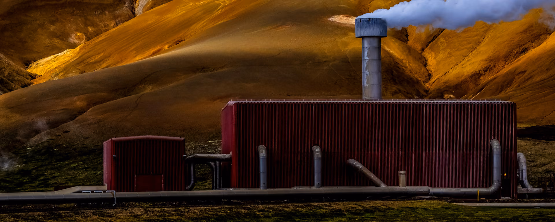 A facility located in hilly terrain with lots of grey pipes going into the walls and a large chimney releasing smoke from the top