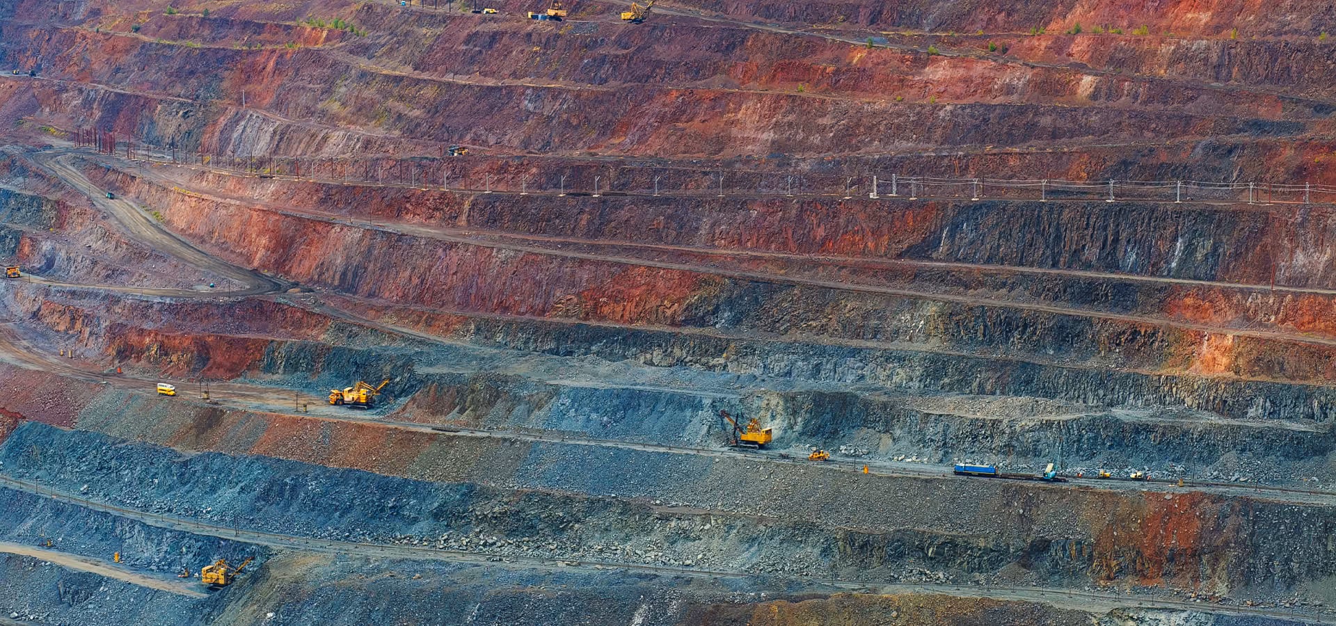 Large open-pit mine with terraced rock layers in varying shades of red, brown, and gray, with yellow mining machinery and trucks on the dirt roads.