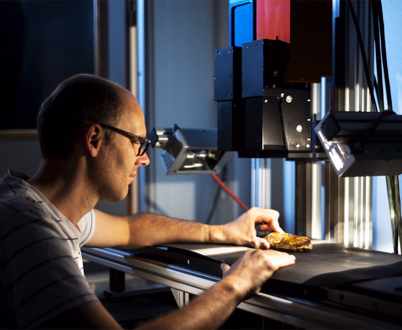 A man with glasses on sitting in a dimly lit room, holding a small mineral piece on a scanning stage underneath two HySpex hyperspectral cameras, one red and one blue