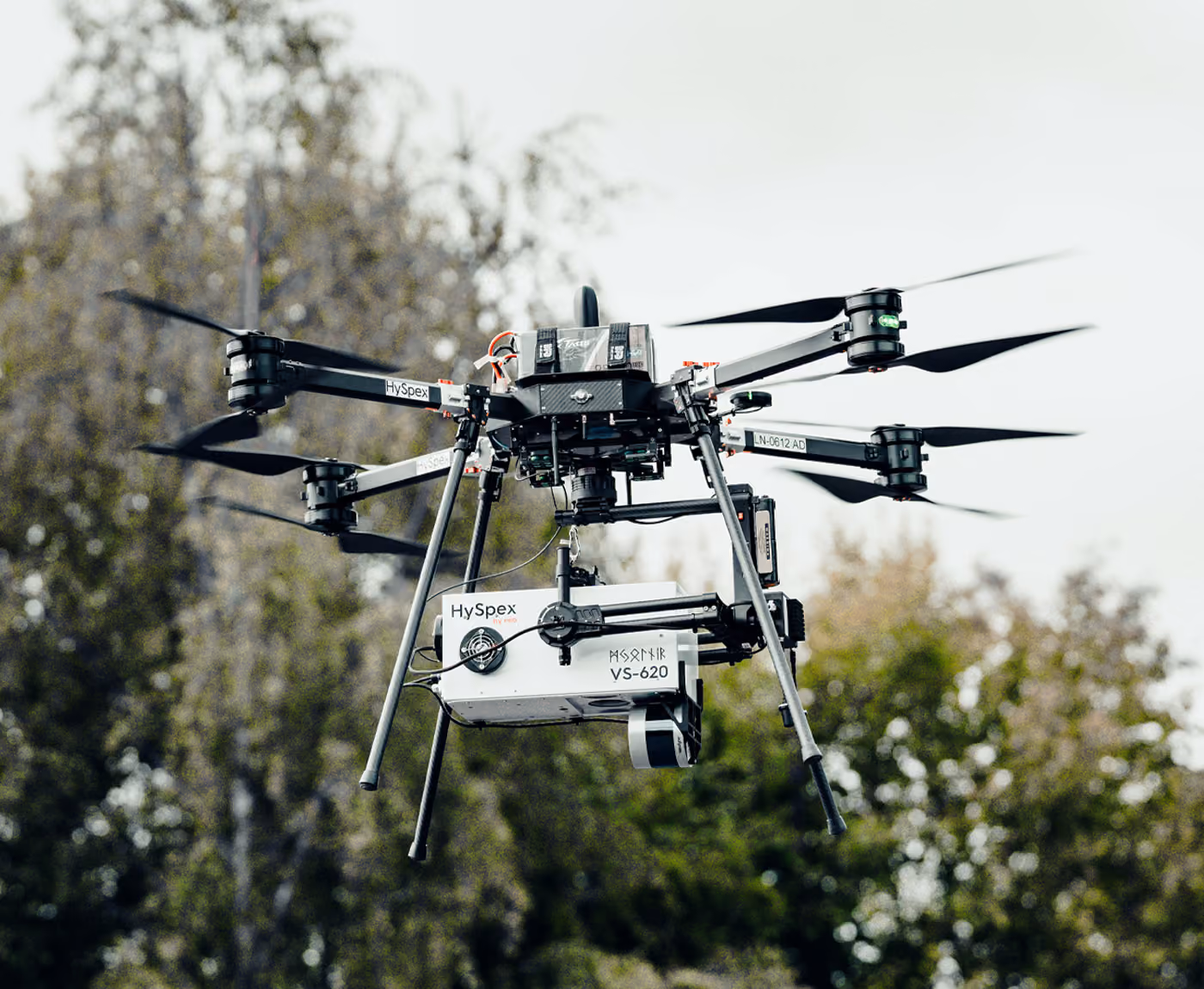 A UAV drone holding a Mjolnir hyperspectral camera while flying through the air with a forest in the background