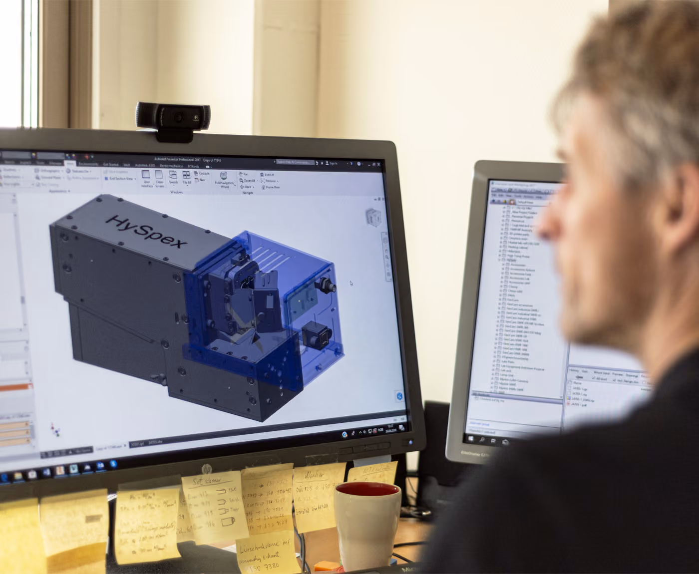 A man with gray hair sitting in front of a PC screen displaying a technical drawing of a blue hyperspectral camera
