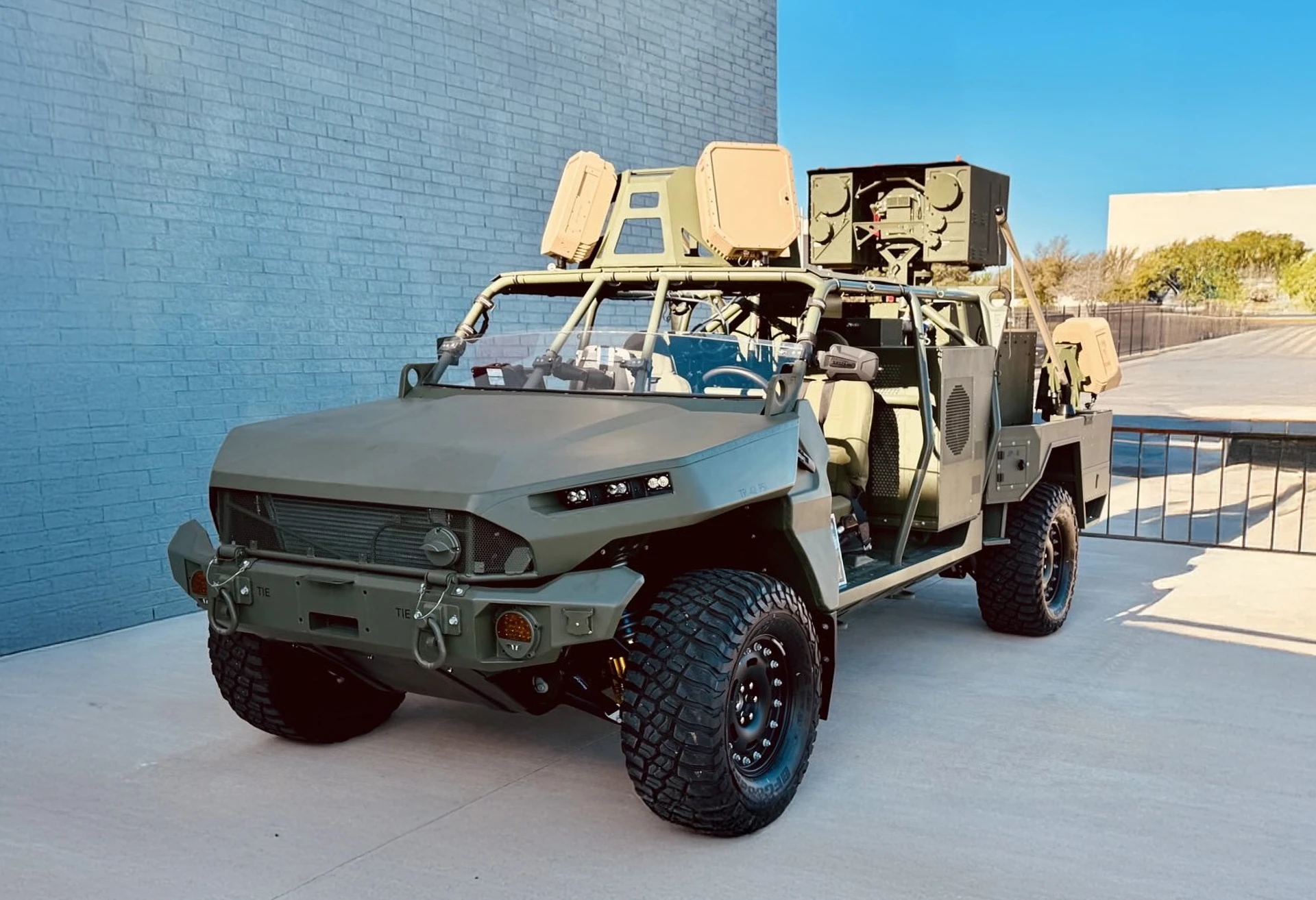 Military light tactical vehicle with mounted EchoShield equipment parked on a concrete surface near a blue brick wall.
