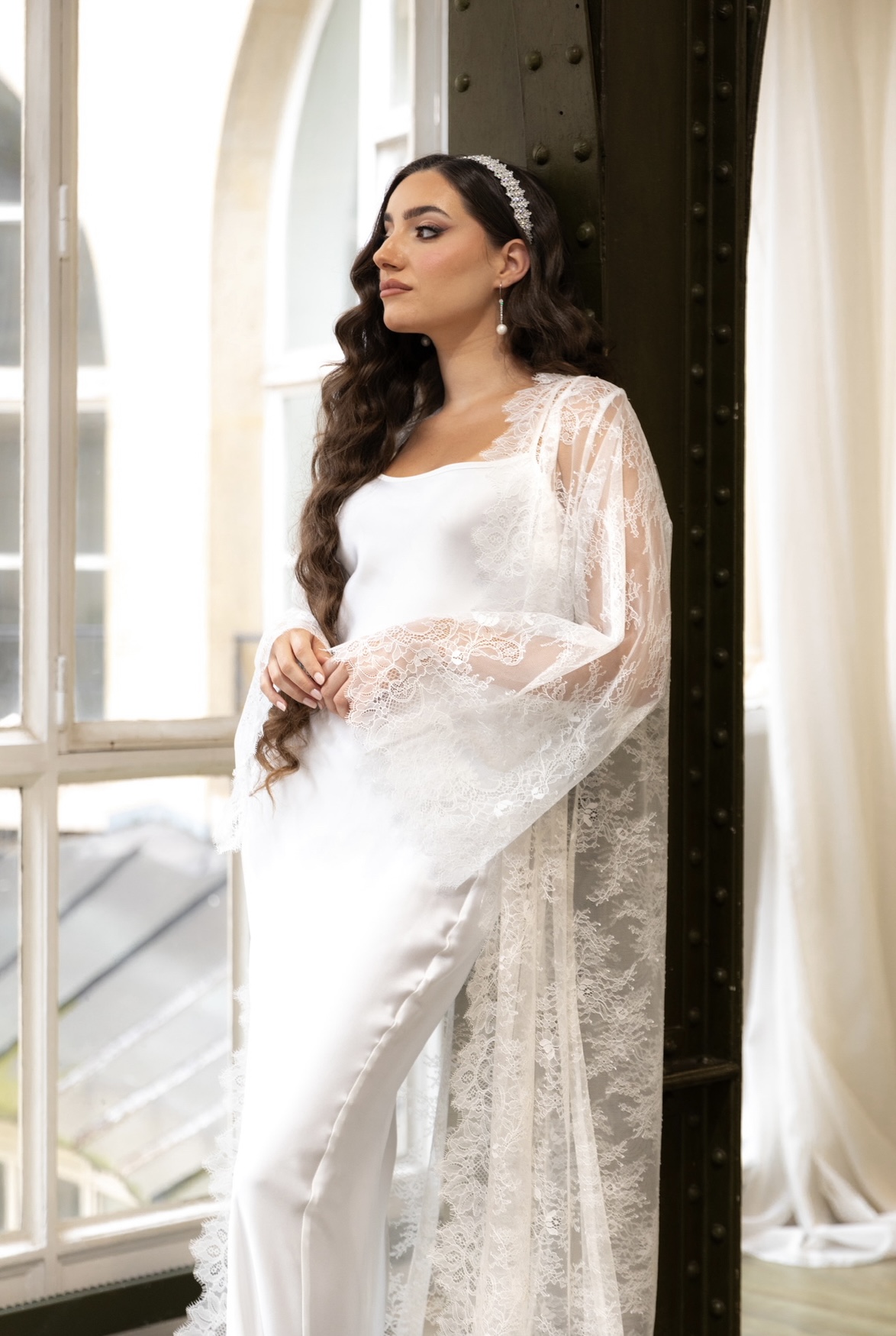 Une mariée portant une robe blanche satinée avec une cape fluide, des sandales blanches et un serre-tête décoré dans une pièce élégante au sol en bois.