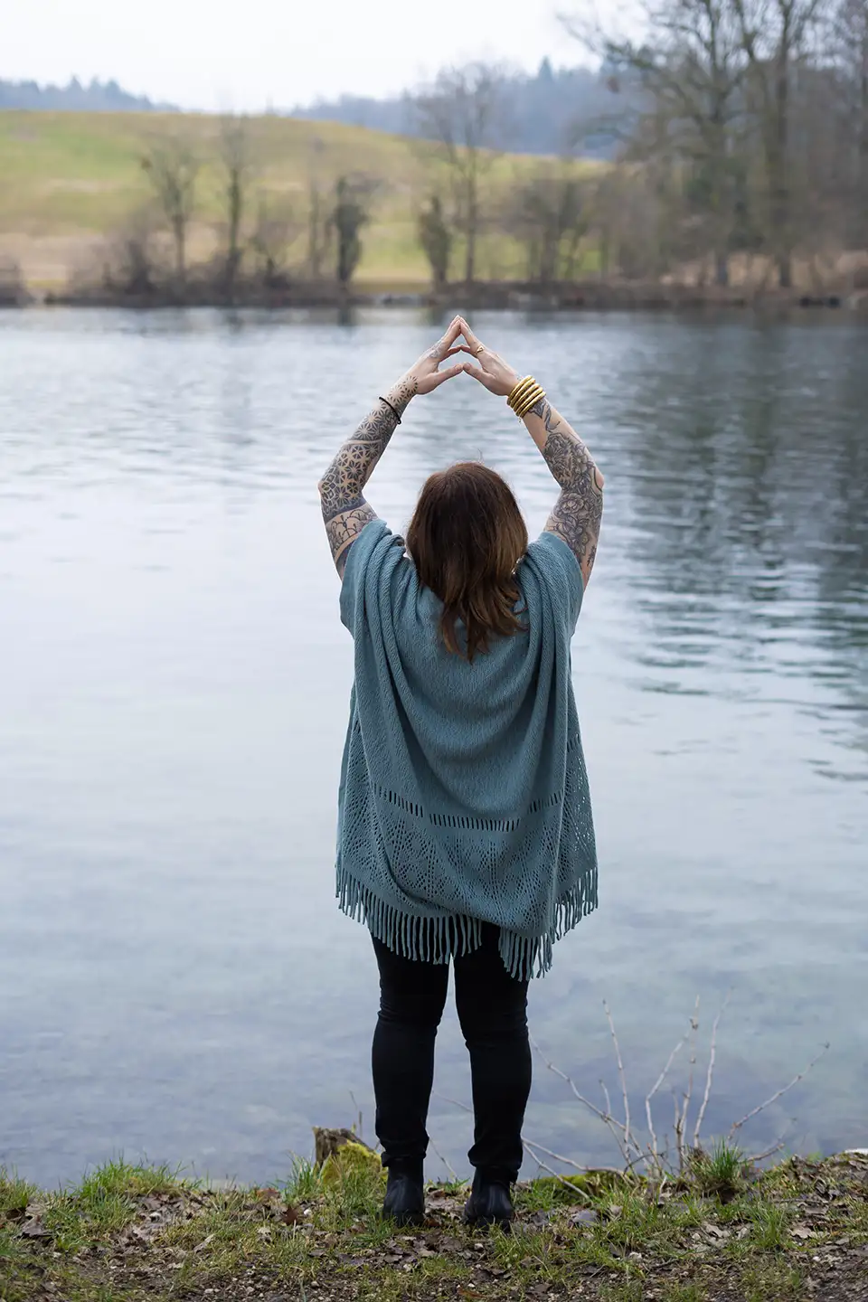 Person with tattooed arms and bracelets standing by a lake, raising hands above head in a triangle shape.