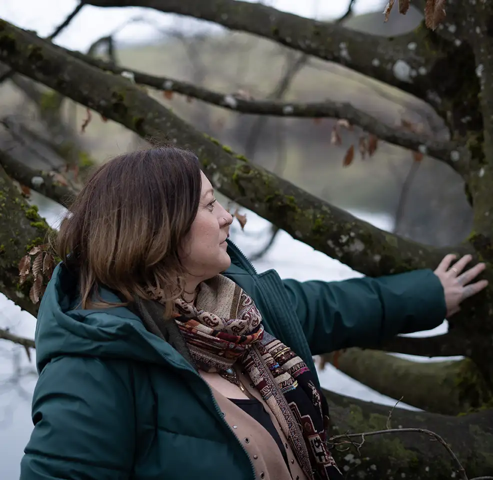 Woman in a green jacket and patterned scarf touching a mossy tree branch near water.