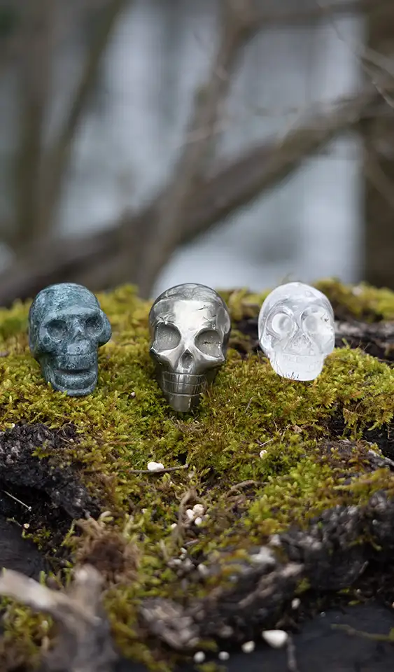 Three small skull-shaped crystals in green, clear, and white standing on moss-covered ground outdoors.