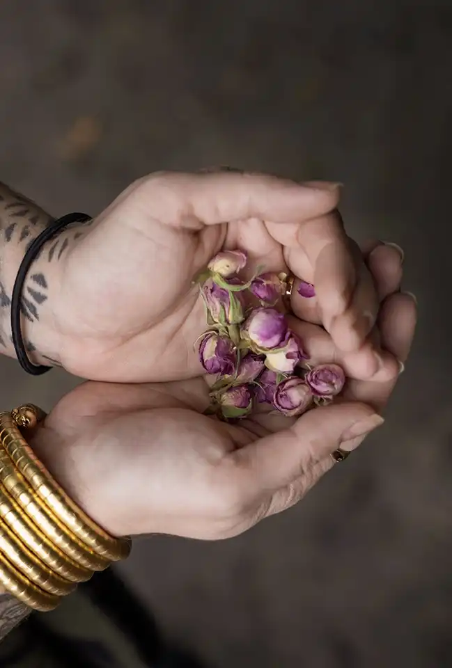 Hands with tattoos and gold bangles holding a small bunch of pink dried rosebuds.