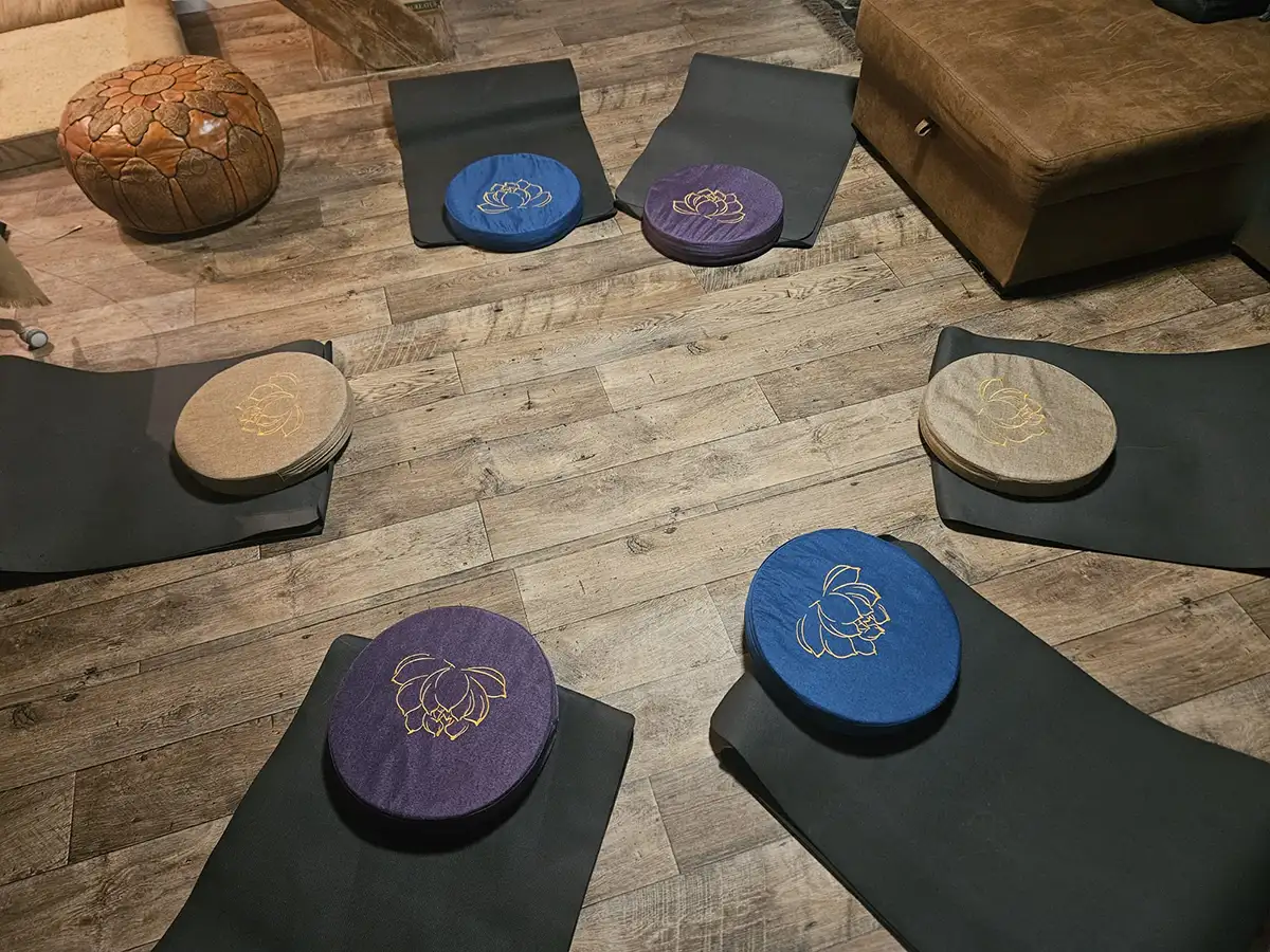 Meditationskissen in Blau, Lila und Grau auf Yoga-Matten in einem Raum mit Holzfußboden, neben einem braunen Sitzpuff.