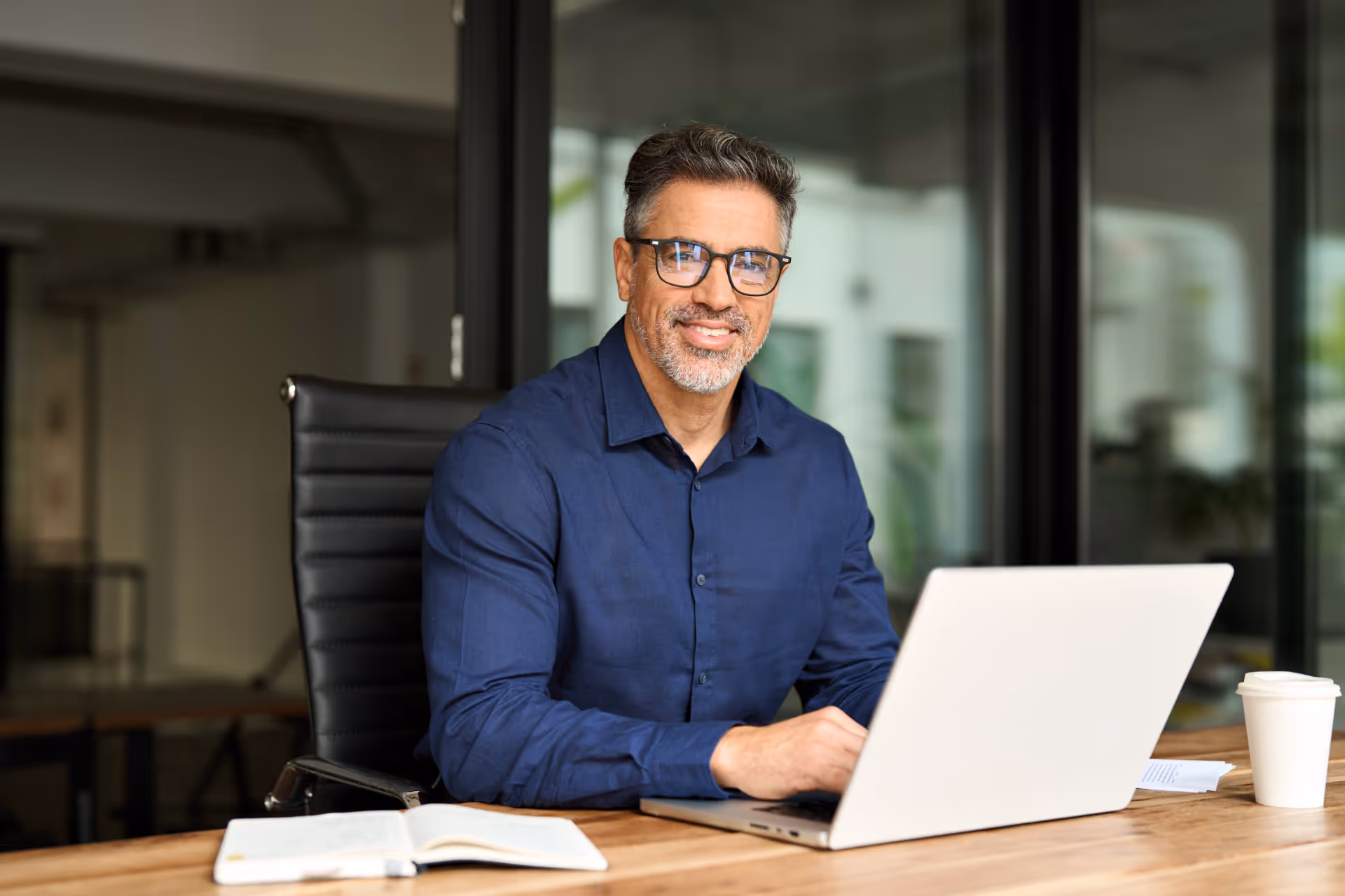 Homme portant des lunettes et une chemise bleue assis à un bureau avec un ordinateur portable, un carnet ouvert et un gobelet en papier.