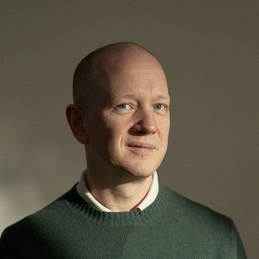 Portrait of a bald man with light skin and blue eyes wearing a green sweater over a white collared shirt against a plain background.