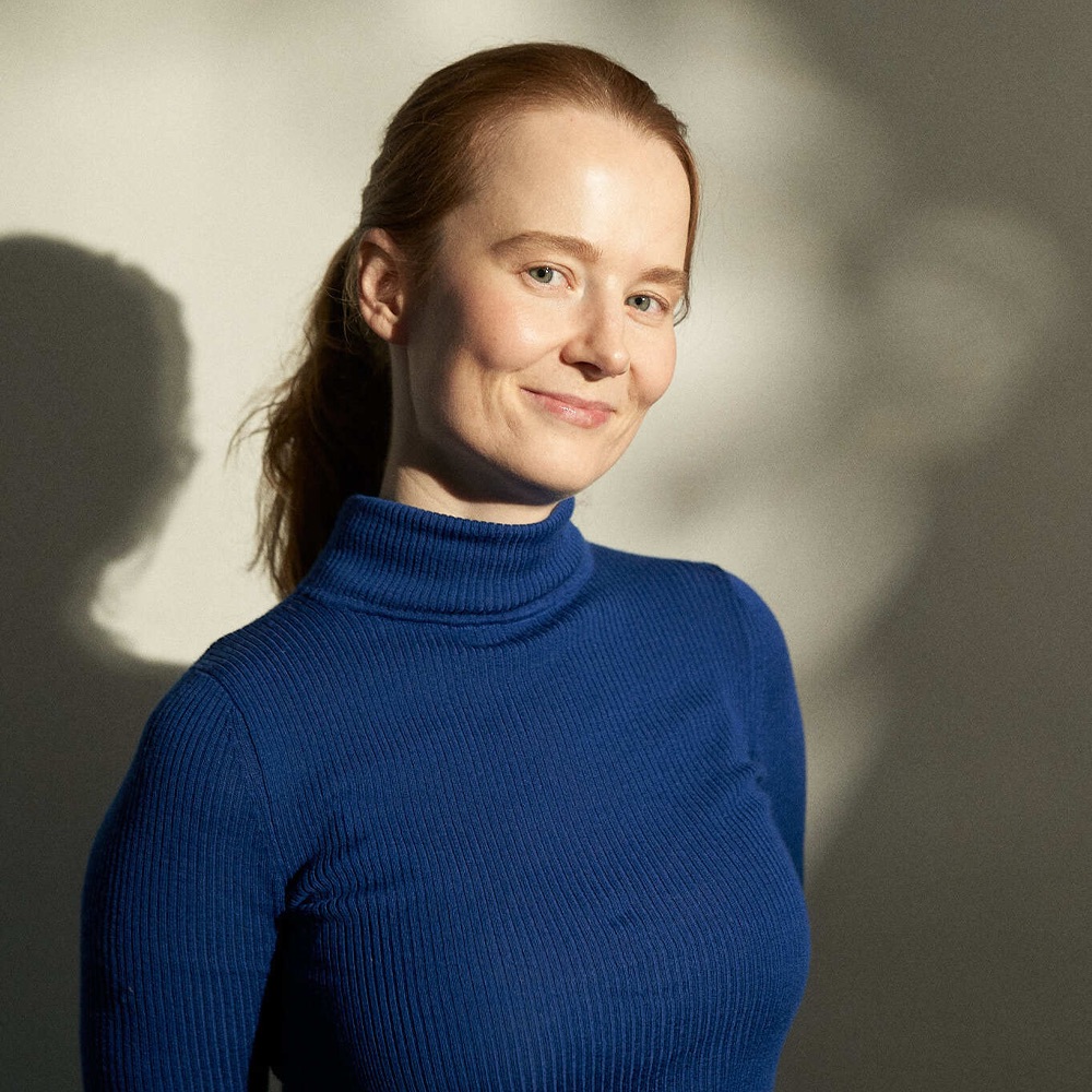 Smiling woman with light skin and red hair tied back wearing a blue ribbed turtleneck sweater against a neutral background.