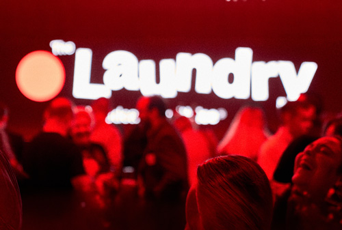 People socializing in a dimly lit venue with a large illuminated 'Laundry' sign in the background.