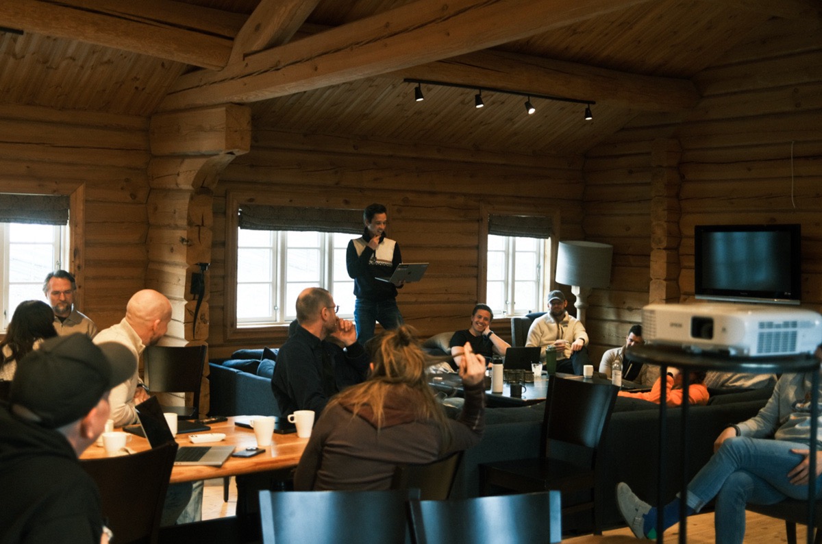Group of people sitting and listening to a man standing and speaking in a cozy wooden room with large windows and a projector.