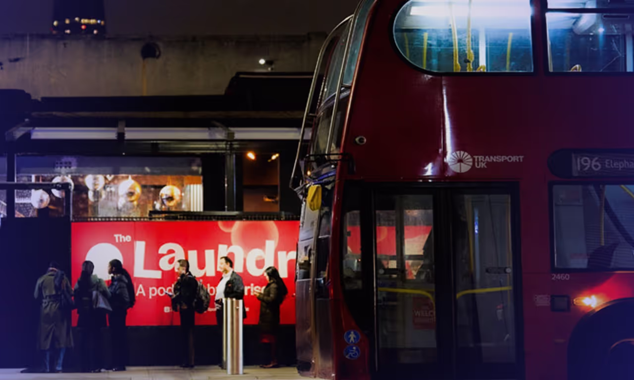 People wait in line at night near a red London double-decker bus in front of a bright red advertisement for The Laundry podcast.