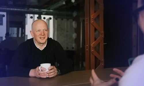 Man in black sweater holding a white mug, engaged in conversation with another person whose hands are visible.