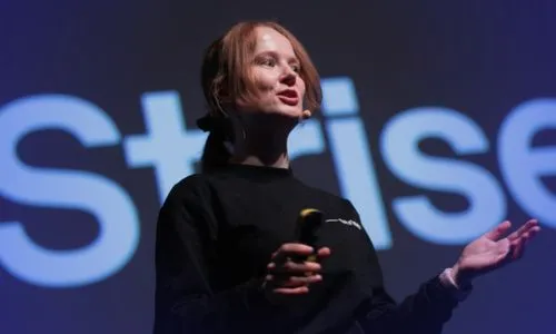 Woman with red hair speaking on stage holding a remote, with a blurred background text starting with 'Strise'.
