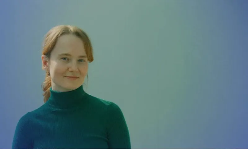 Woman with light skin and red hair braided, wearing a dark green turtleneck sweater, smiling softly against a gradient blue-green background.
