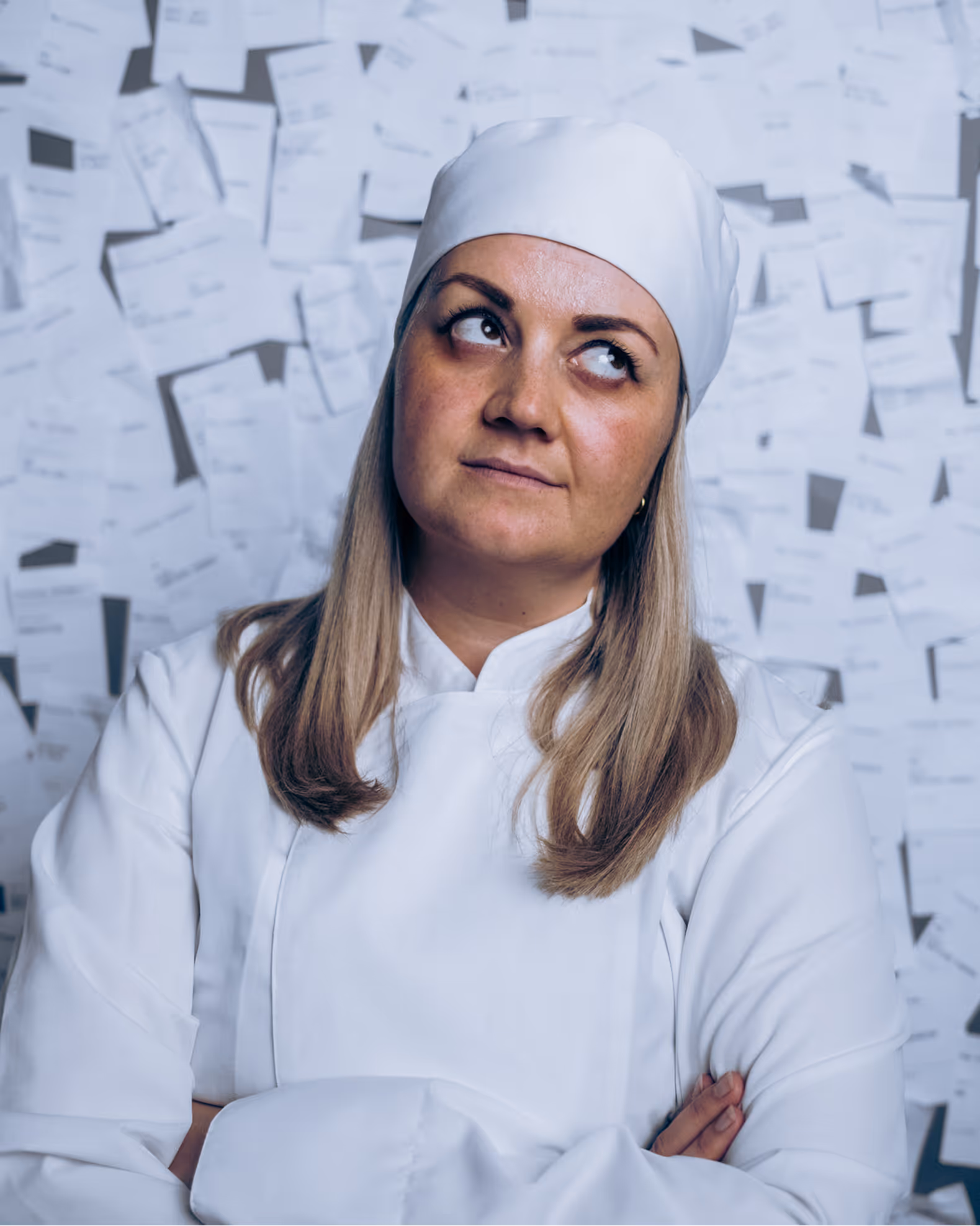 Woman chef with arms crossed wearing white uniform and hat looks up thoughtfully against receipt-covered wall.