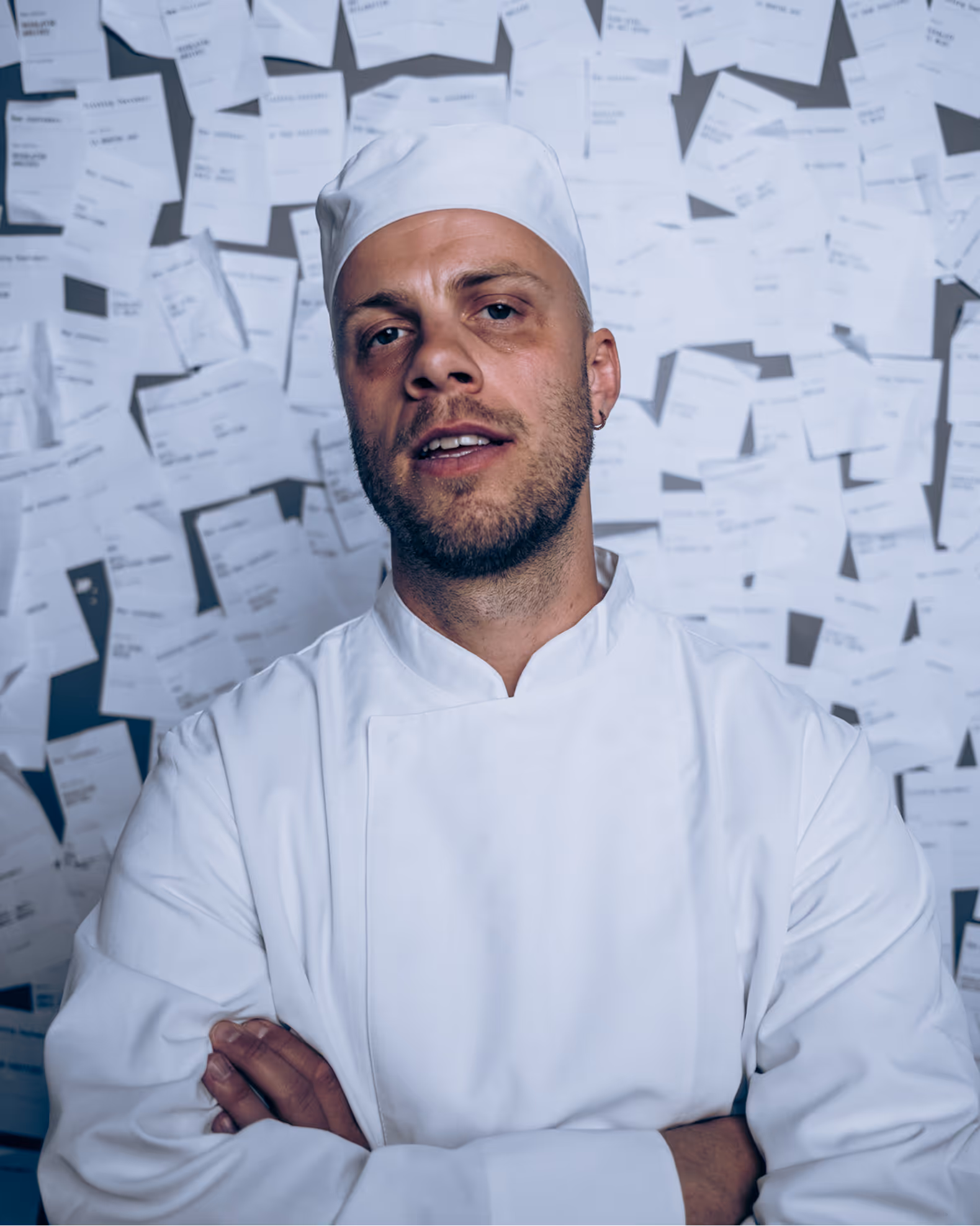 Male chef in white uniform and hat stands with arms crossed against wall covered in papers.