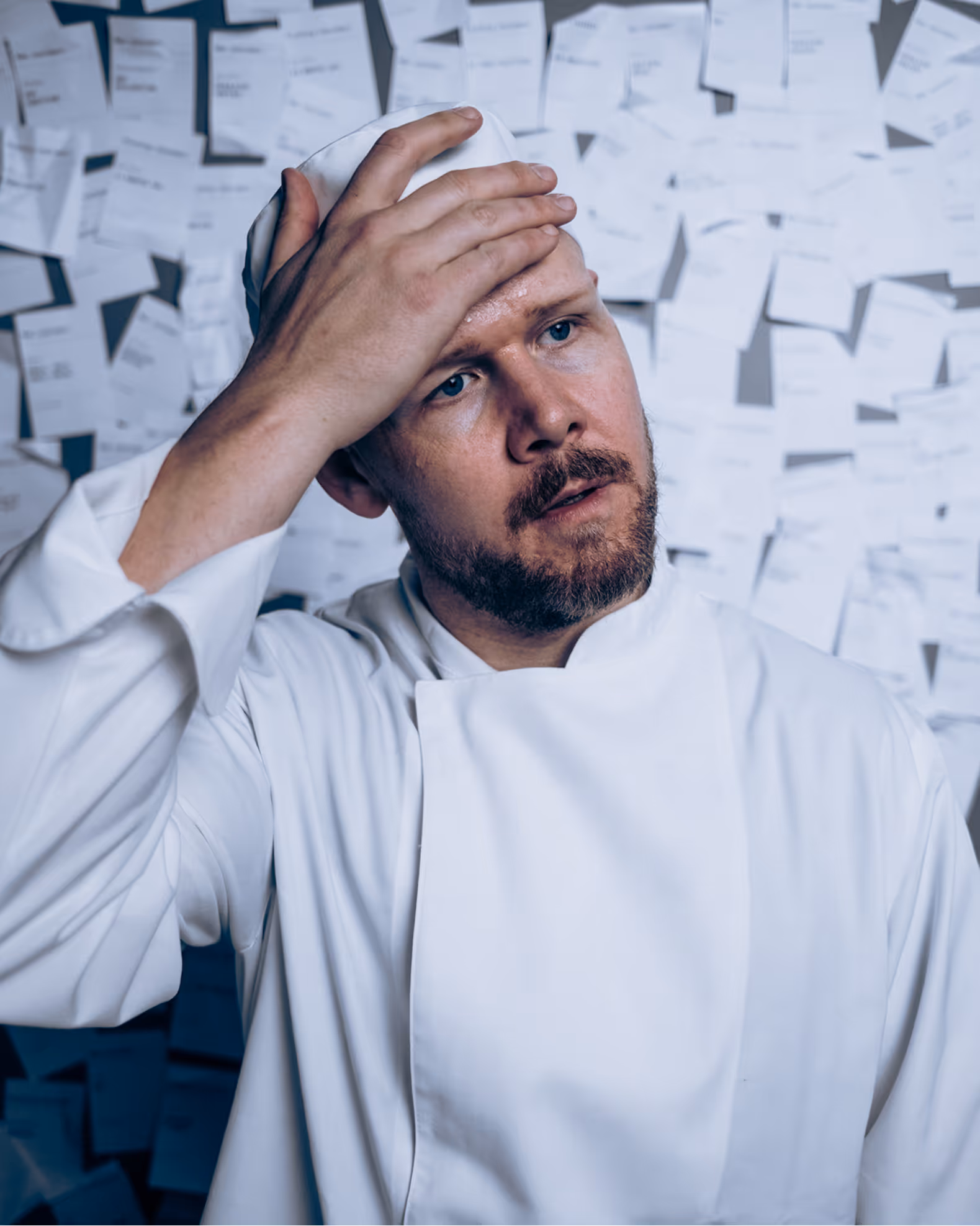Worried chef in white uniform touching his forehead with many papers pinned on the wall behind him.