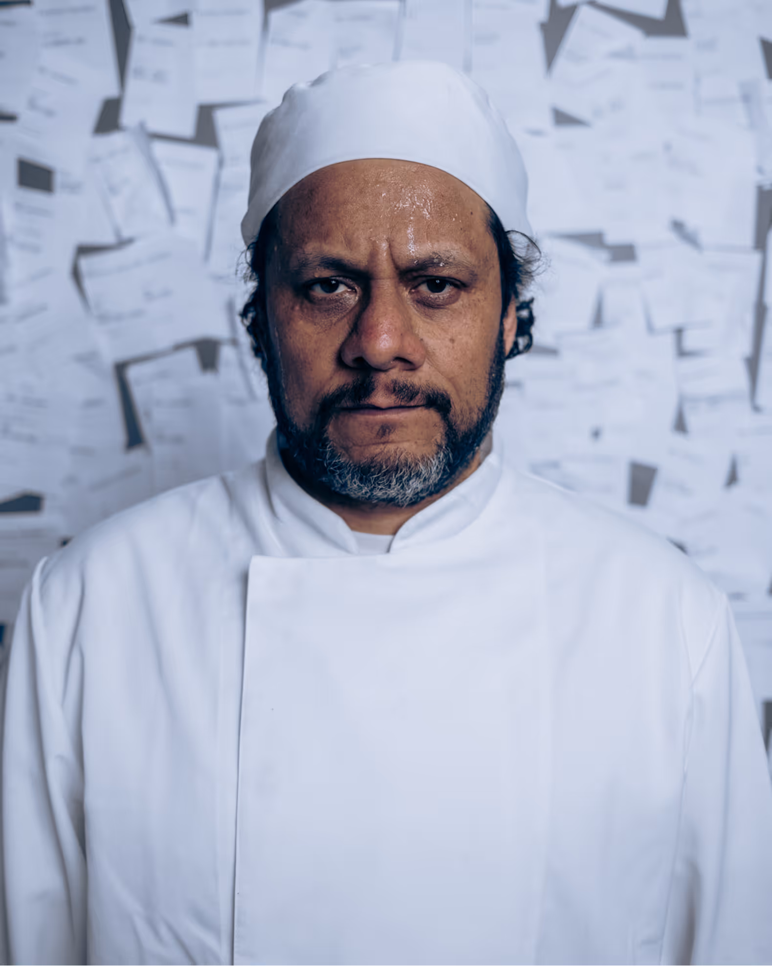 Chef in white uniform and hat stands against a wall covered with pinned papers.