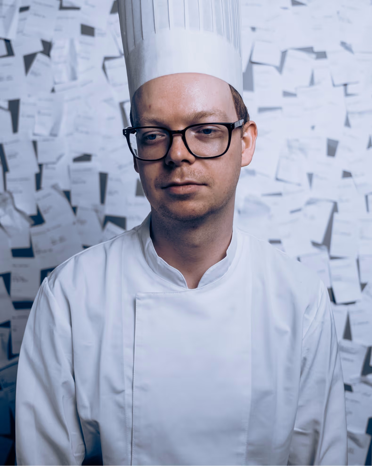 Man in white chef uniform and tall hat standing in front of a wall covered with papers.
