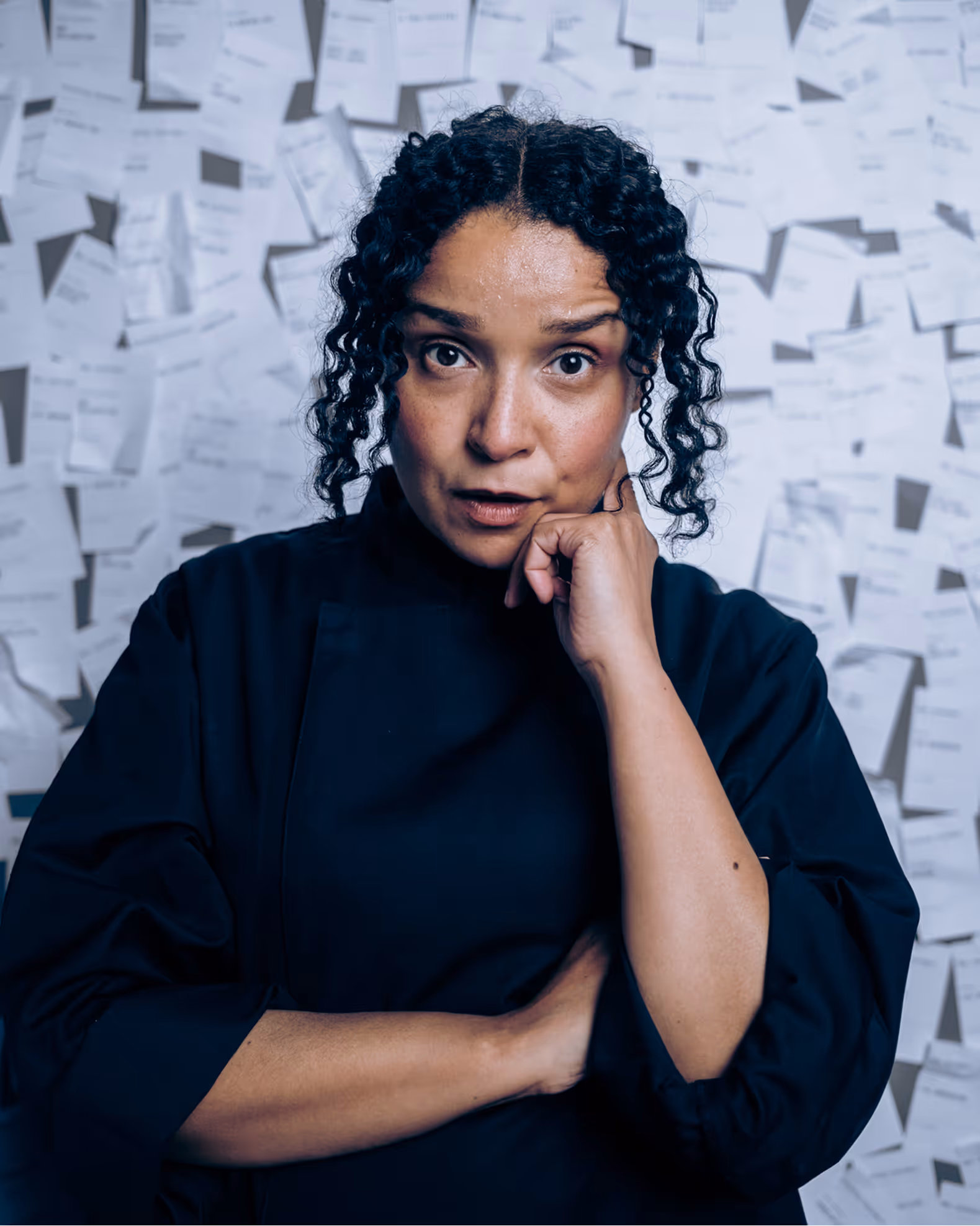 Woman with curly hair in black shirt, thoughtful pose with hand on chin, papers on wall behind her.