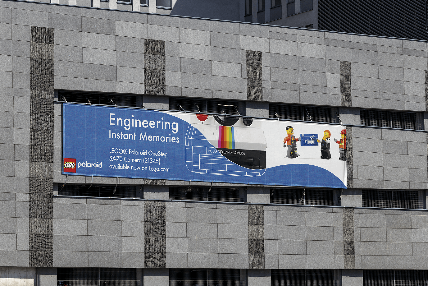 Large banner on grey building wall showing LEGO Polaroid OneStep SX-70 Camera with Lego minifigures holding a blueprint.