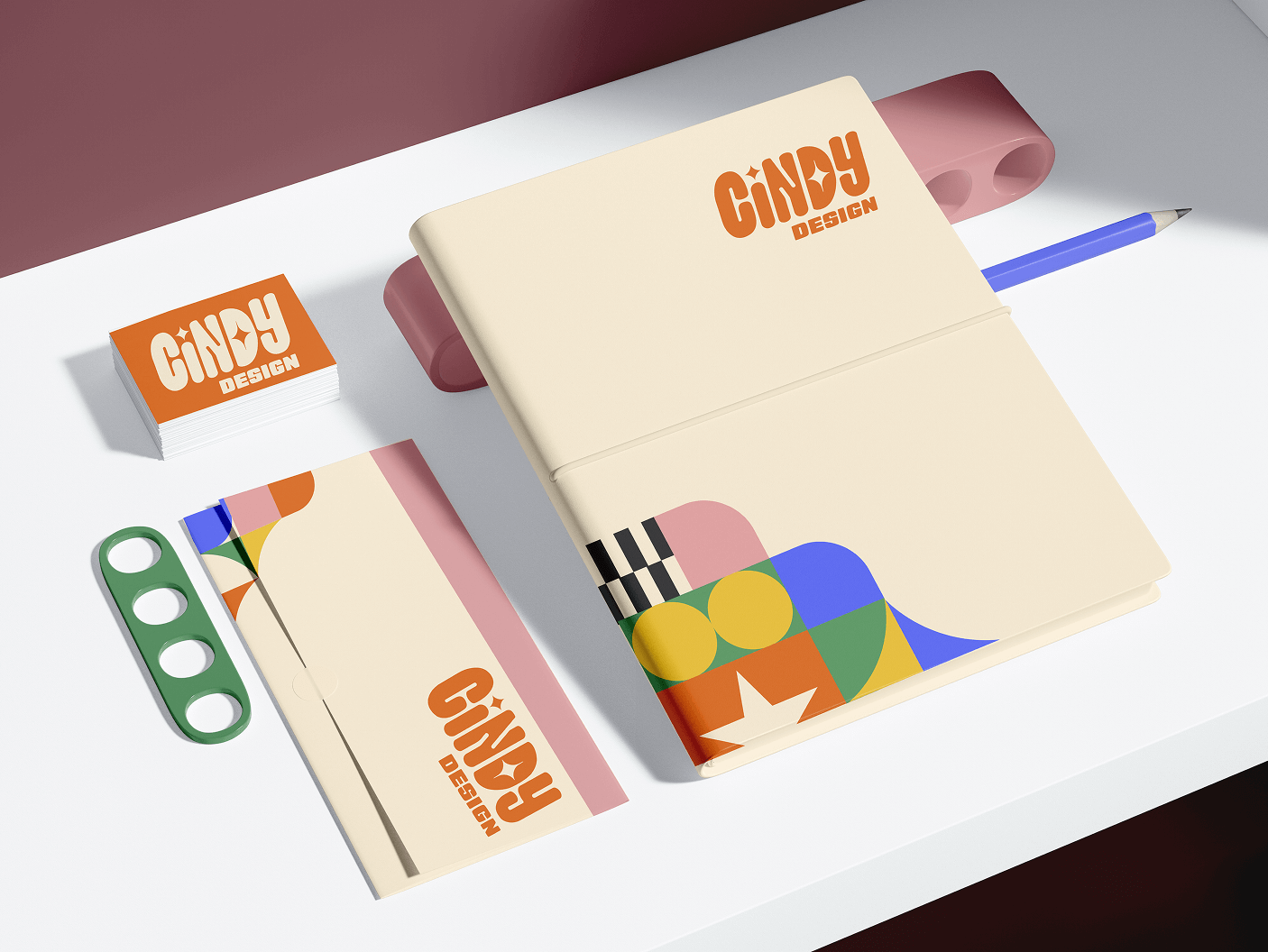 Collection of Cindy Design stationery including notebook, business cards, envelope, green paint palette, and blue pencil on white table.