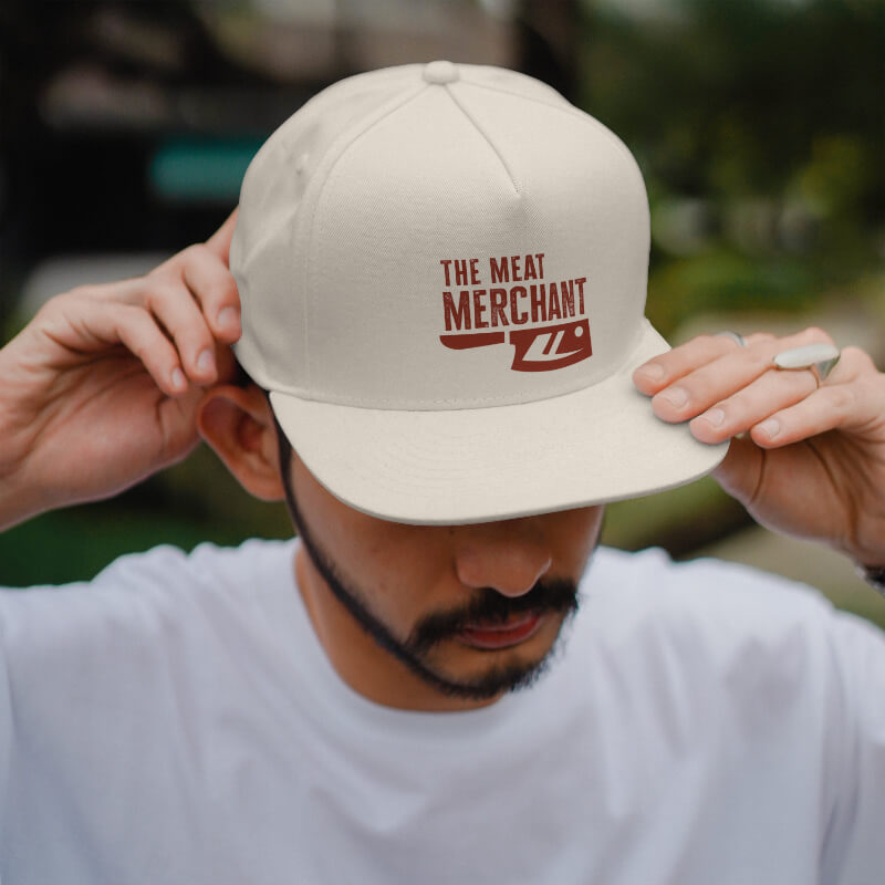 Man wearing a light-colored hat with 'THE MEAT MERCHANT' and a cleaver graphic on the front.