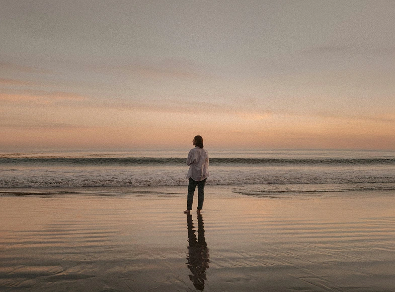 Person standing barefoot on wet sand at the beach, facing the ocean during a calm sunset.