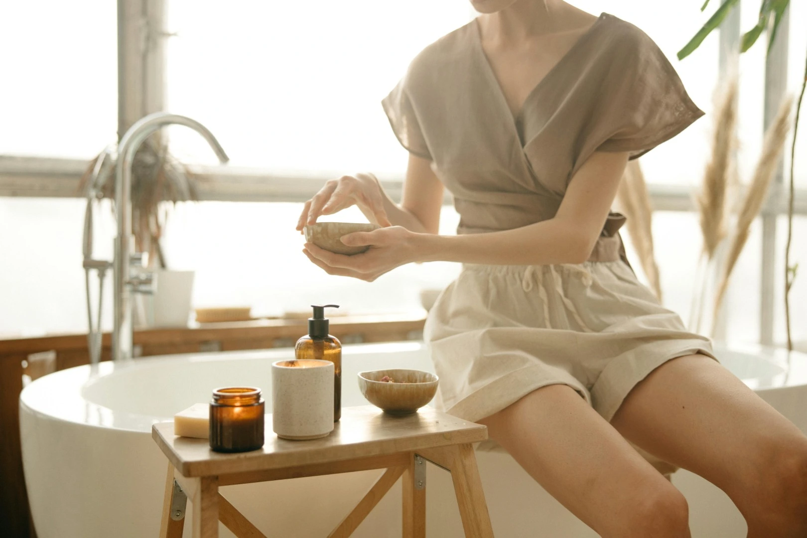 Woman sitting by a bathtub applying skincare product from a small bowl with spa items on a wooden stool nearby.