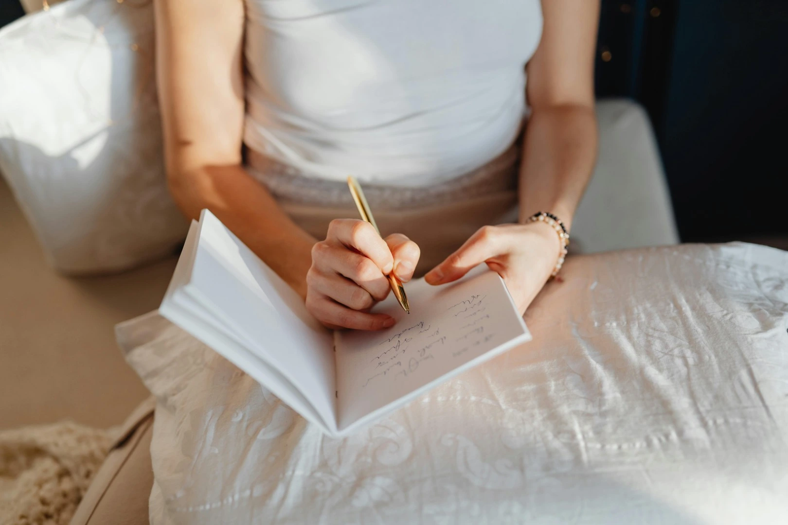 Person sitting on a bed writing with a gold pen in a notebook resting on a white quilt.
