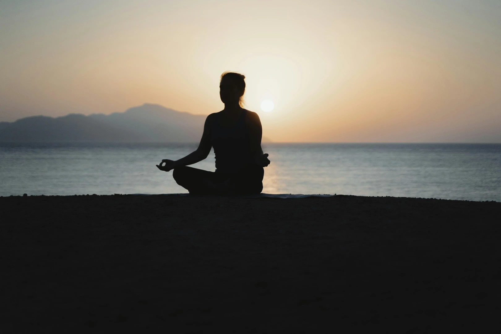 Silhouette of a person meditating in lotus pose on the beach at sunset with mountains in the background.