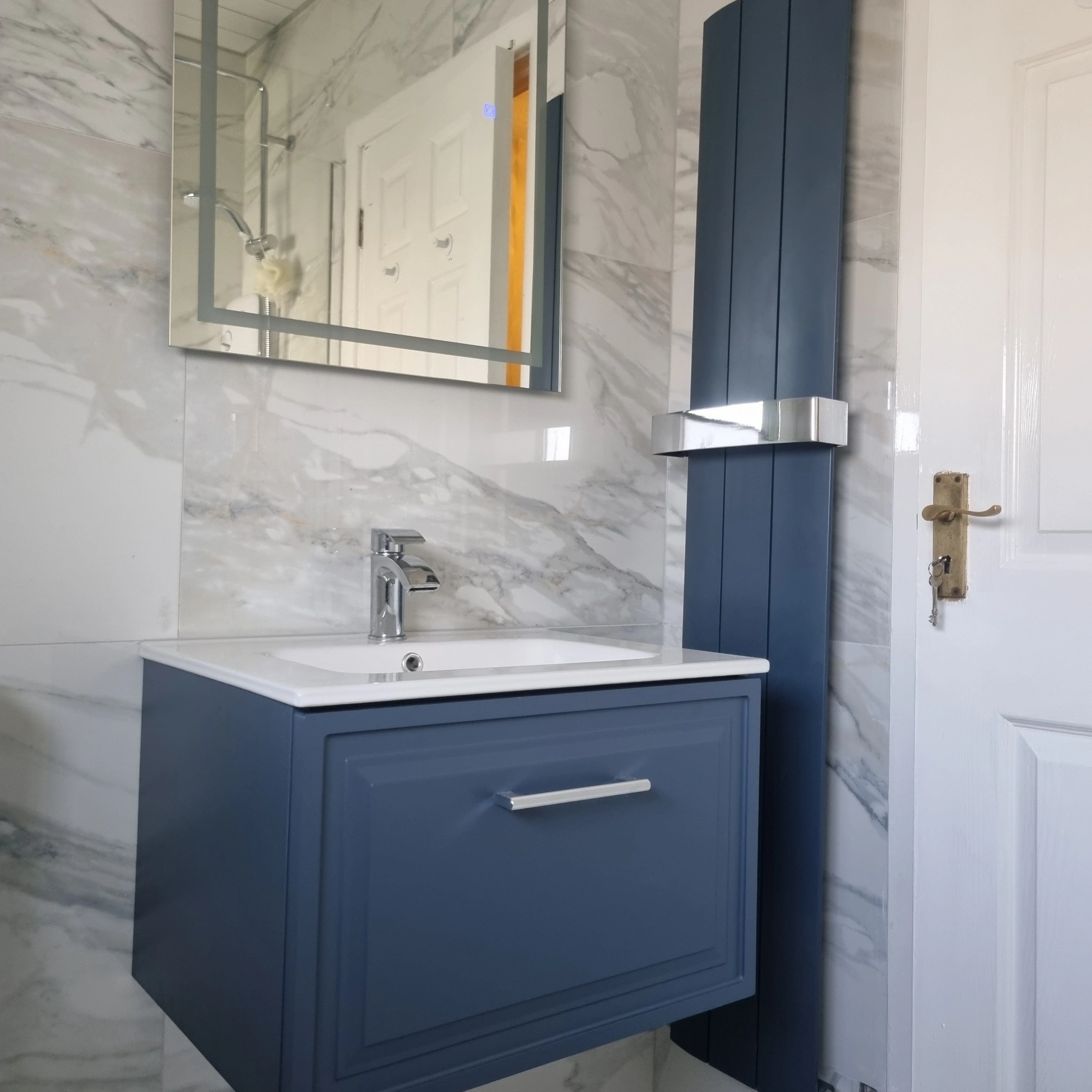 Modern bathroom with a blue floating vanity unit, white sink, chrome faucet, a mirrored cabinet, and marble-patterned wall tiles.