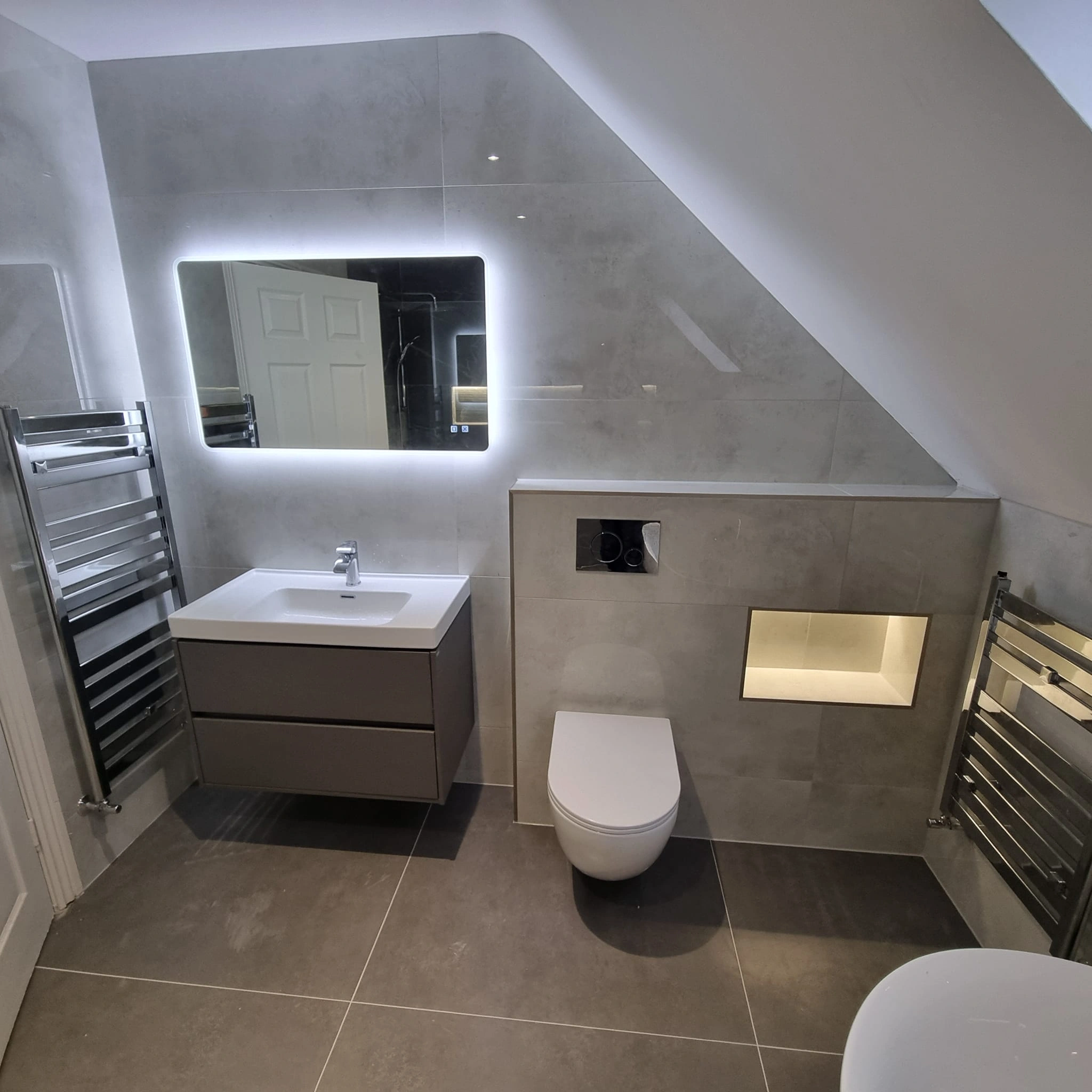 Modern bathroom with wall-mounted white toilet, floating vanity with sink, illuminated rectangular mirror, and chrome heated towel rails.