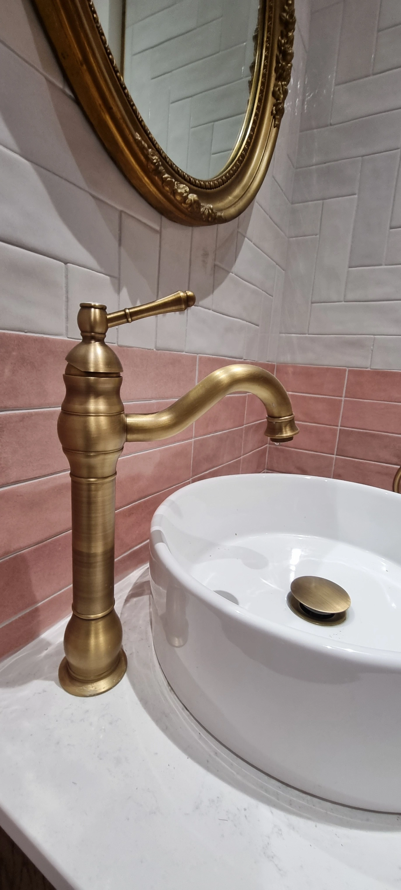 Modern bathroom sink setup with a tall brushed brass faucet, white round basin, and marble countertop with pink and white tiled walls.