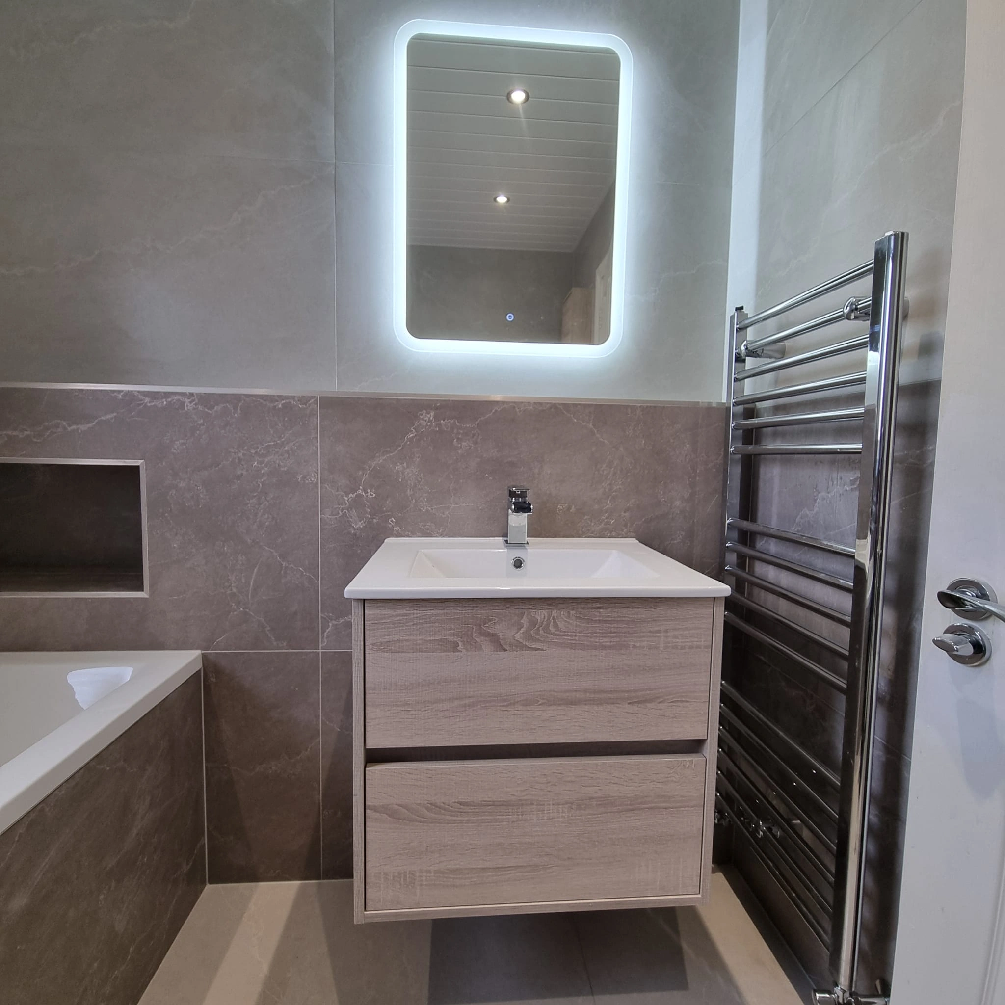 Modern bathroom with a wall-mounted wooden vanity, illuminated rectangular mirror, chrome towel rack, and bathtub with marble tiles.