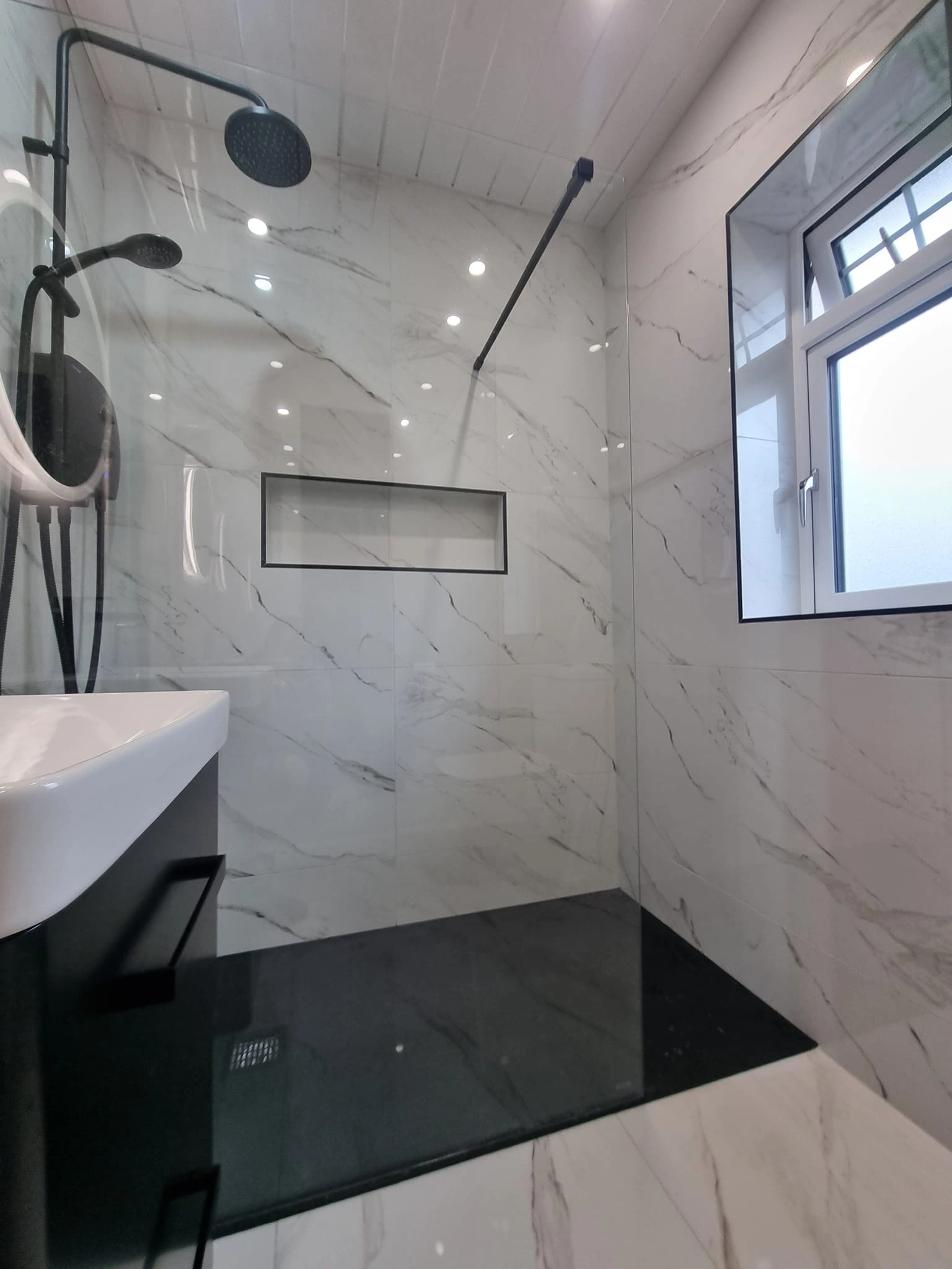 Modern bathroom shower area with black shower fixtures, black shower base, white marble walls, and a window.