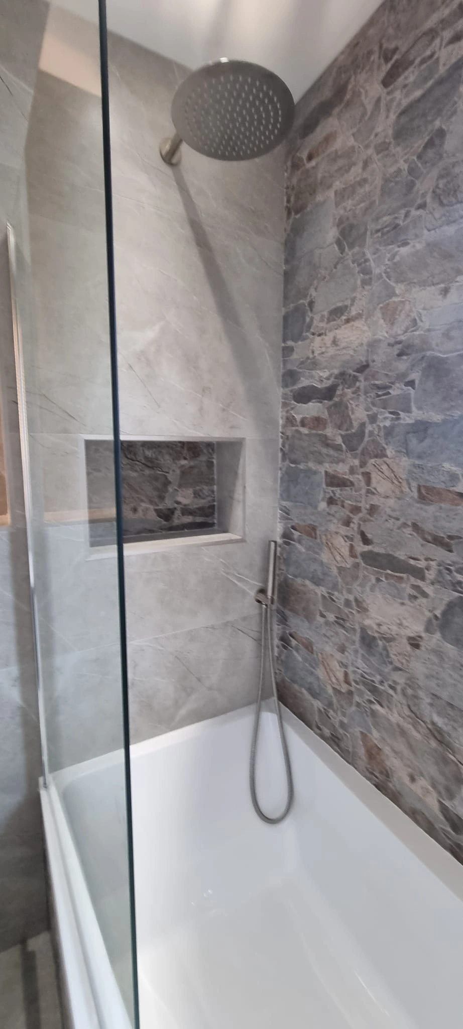 Modern shower with a round overhead showerhead, handheld sprayer, and stone-textured wall tiles inside a white bathtub.