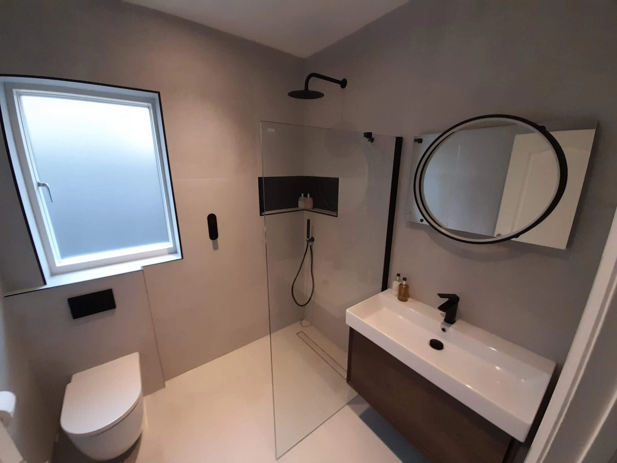 Modern bathroom with a glass walk-in shower, wall-mounted toilet, rectangular sink with black faucet, and round mirror.