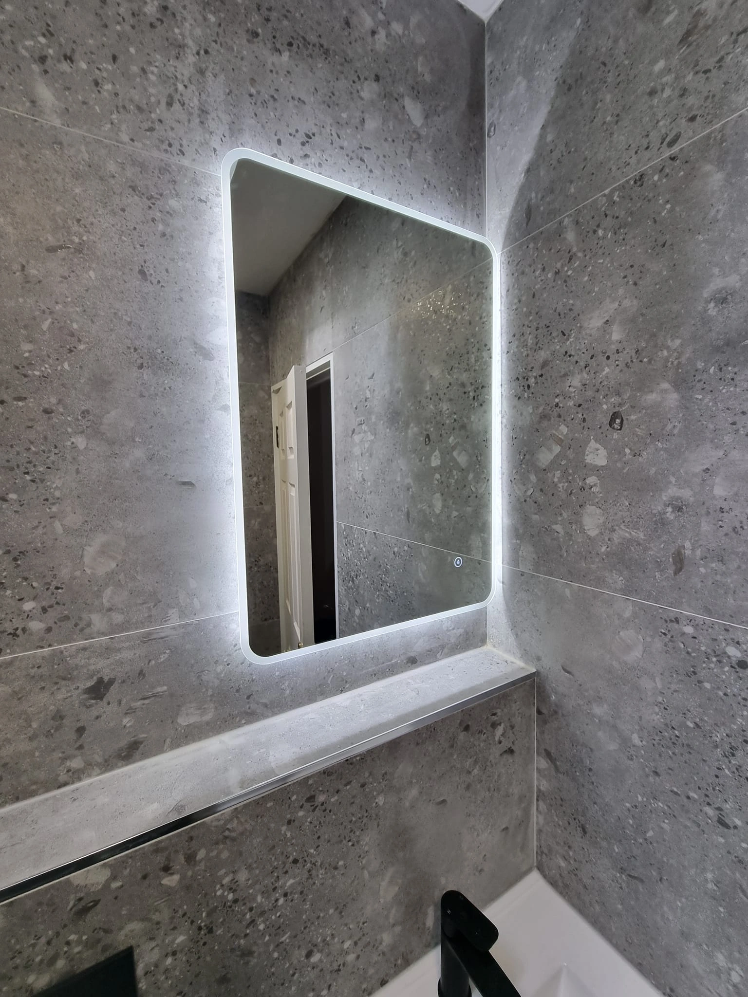 Rectangular bathroom mirror with rounded corners and backlit LED mounted on a gray speckled stone tiled wall above a white sink with a black faucet.